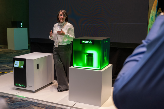 A woman standing between two gas generators - one a white cuboid with a black front panel and tilting display screen, the other a concept box of frosted glass with a green glow coming from inside. This concept box is showing components that are inside the generator.